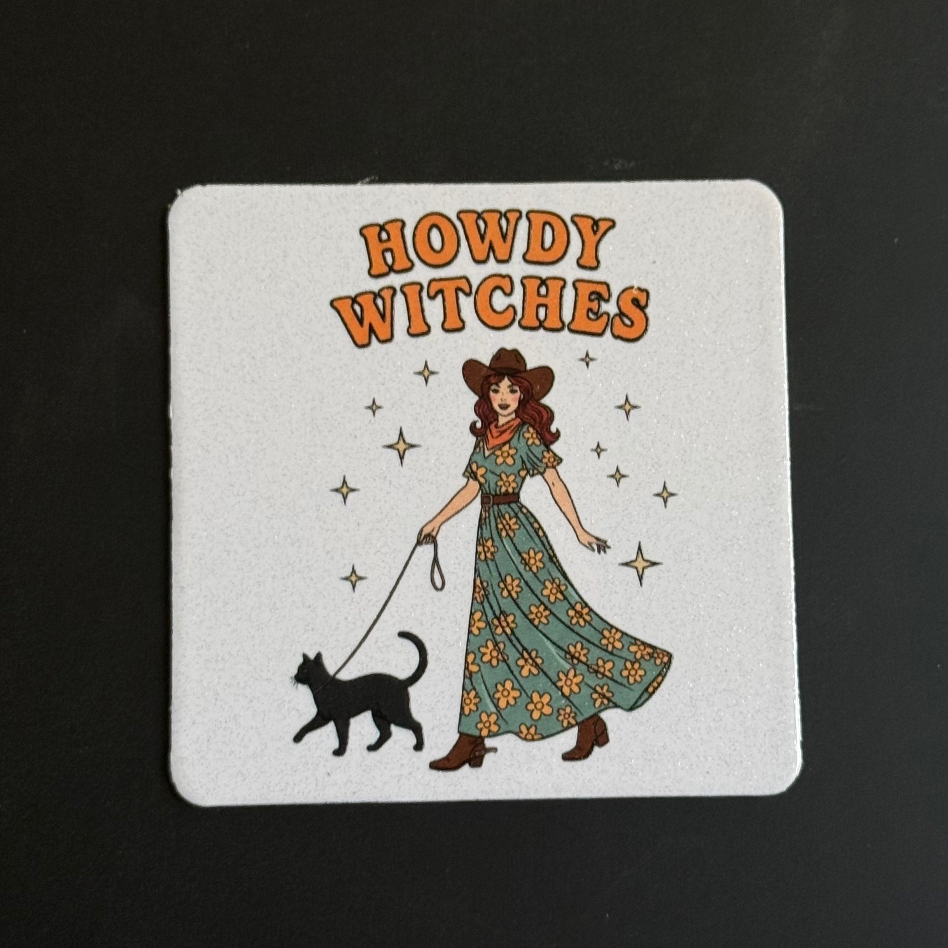 A square white coaster with rounded corners featuring a retro illustration of a woman in a long floral dress walking a black cat, with the words 'HOWDY WITCHES' in orange above. The coaster is photographed on a dark black background.