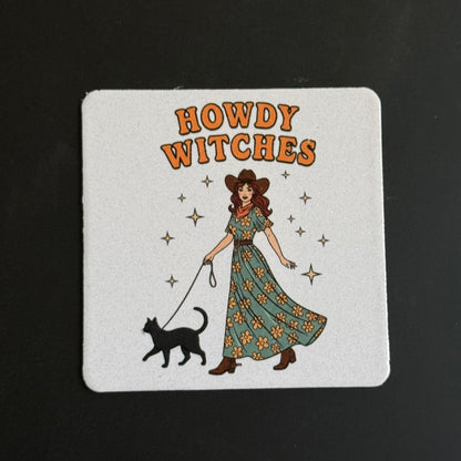 A square white coaster with rounded corners featuring a retro illustration of a woman in a long floral dress walking a black cat, with the words 'HOWDY WITCHES' in orange above. The coaster is photographed on a dark black background.
