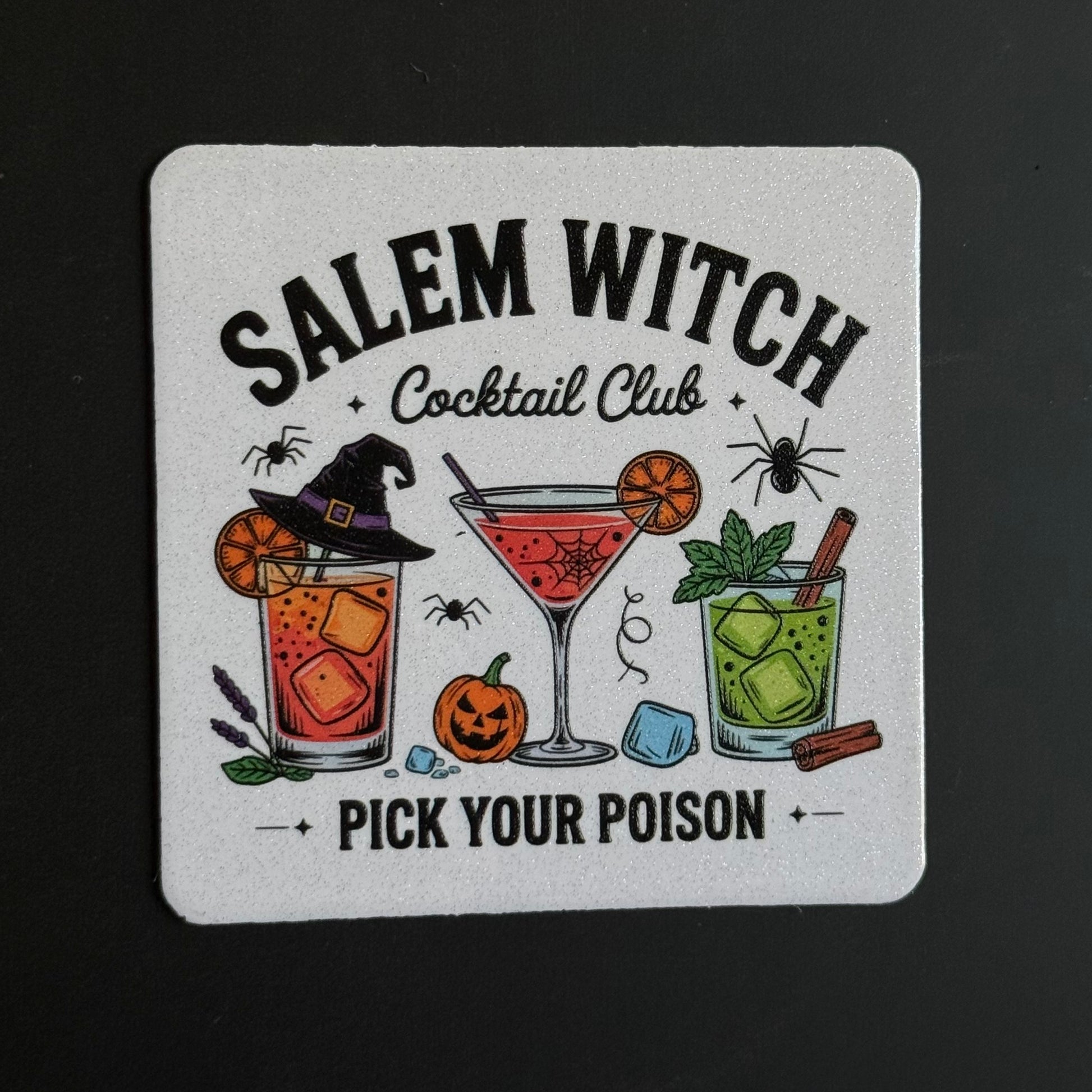 A square white coaster with a Halloween-themed design reading 'SALEM WITCH Cocktail Club' and 'PICK YOUR POISON'. The coaster features illustrations of three cocktails (orange, pink, and green), a witch hat, spiders, a pumpkin, and a cinnamon stick on a dark background.
