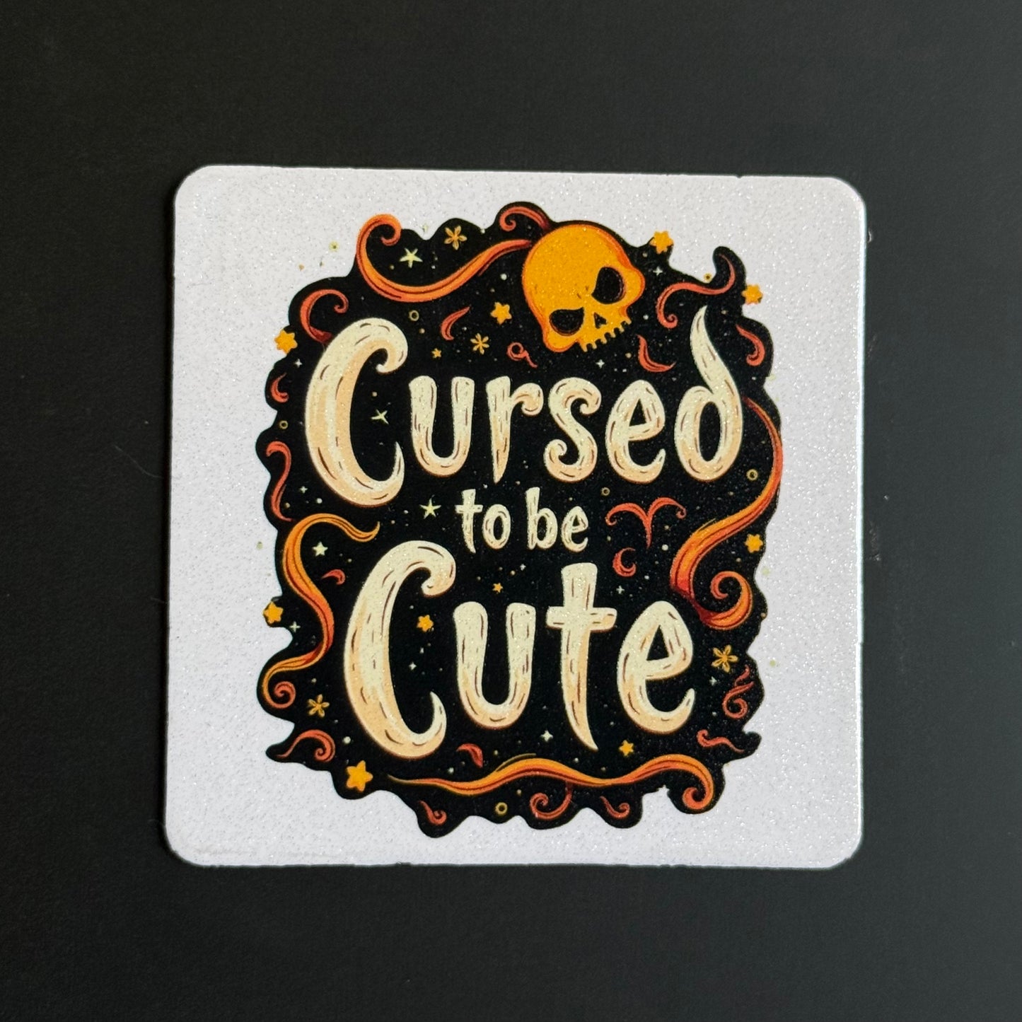A square white coaster featuring a Halloween-themed illustration with the phrase 'Cursed to be Cute' in cream lettering, surrounded by orange swirls, stars, and a small orange skull on a black background.