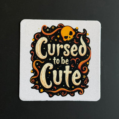 A square white coaster featuring a Halloween-themed illustration with the phrase 'Cursed to be Cute' in cream lettering, surrounded by orange swirls, stars, and a small orange skull on a black background.