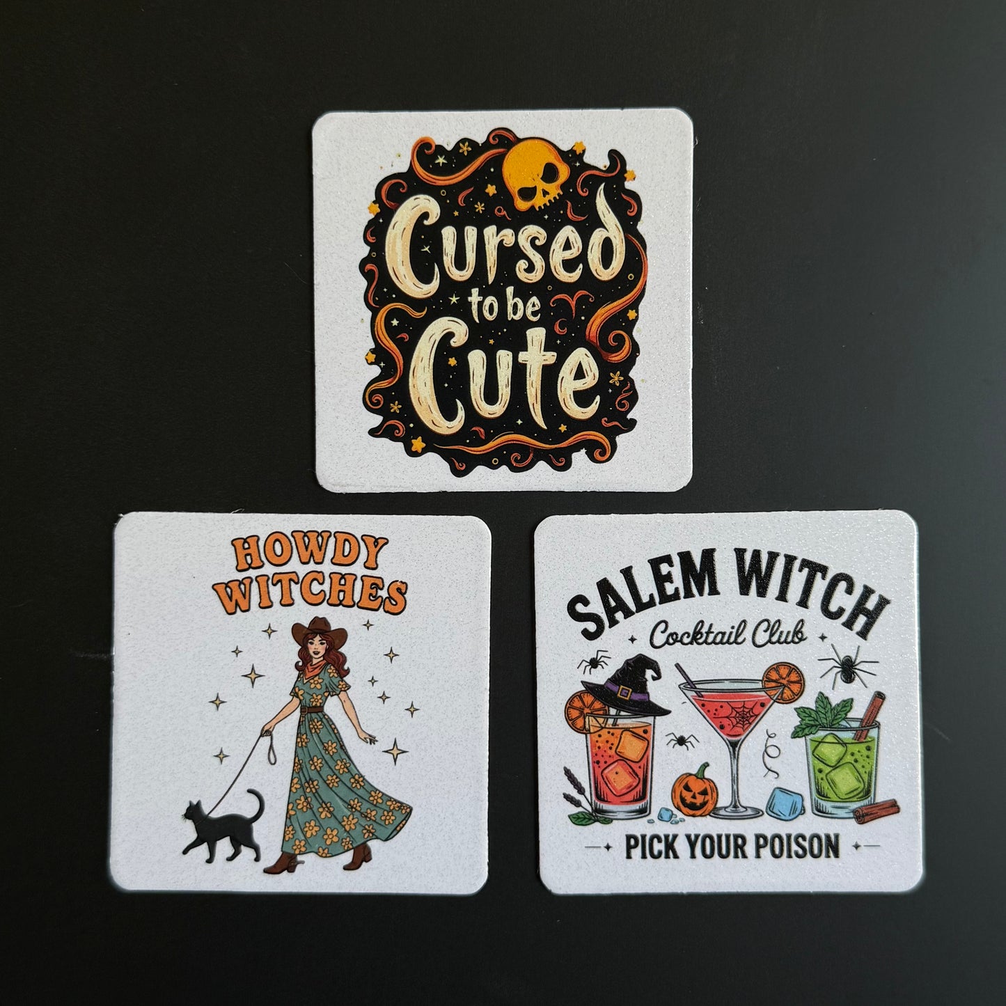 Three square white coasters with Halloween-themed illustrations and text, arranged on a dark background. The top coaster reads 'Cursed to be Cute' with a skull and decorative swirls.