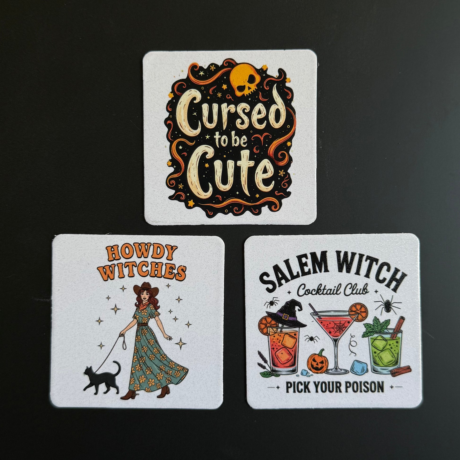 Three square white coasters with Halloween-themed illustrations and text, arranged on a dark background. The top coaster reads 'Cursed to be Cute' with a skull and decorative swirls.