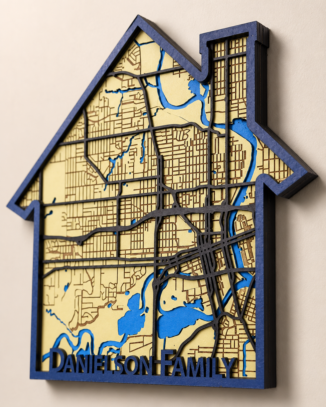 A house-shaped wooden wall plaque featuring a custom map design with layered black street lines, blue waterways, and a cream-colored wood background. The bottom is personalized with the text 'DANIELSON FAMILY' in dark blue.