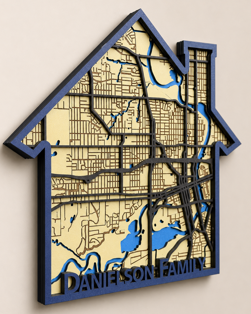 A house-shaped personalized wooden map wall art piece featuring the street grid  with blue waterways and the custom name 'DANIELSON FAMILY' at the bottom, mounted on a beige wall.